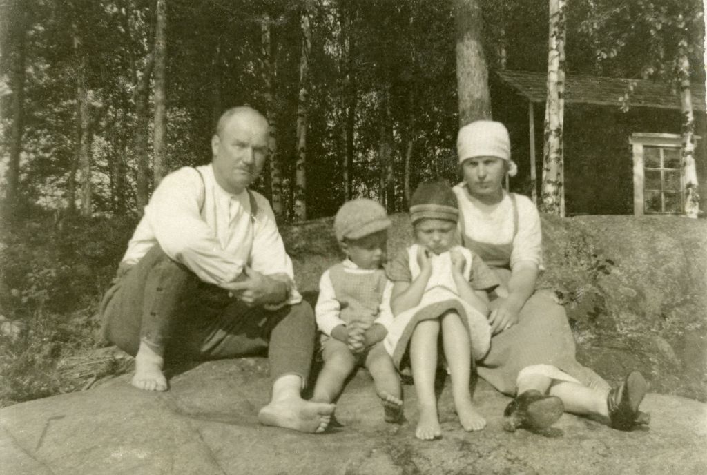 Mies, kaksi lasta ja nainen kalamajan edustalla. Vanha, mustavalkoinen kuva.